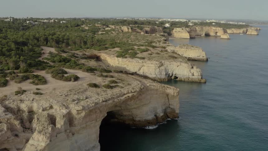 This aerial footage captures a panoramic view of a rugged coastline featuring limestone cliffs and sea caves the camera glides over the rocky landscape revealing a dense green forest inland and the calm blue ocean stretching toward the horizon