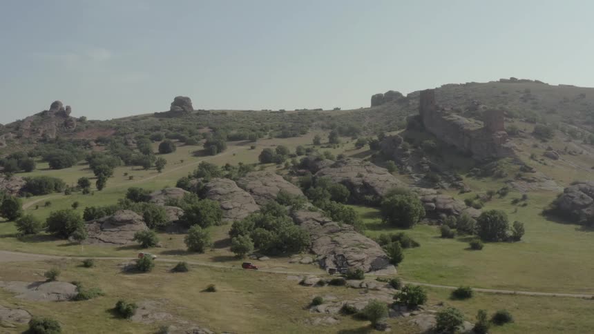 An aerial shot captures three offroad vehicles driving along a winding dirt path through a vast rocky landscape the rugged terrain is dotted with green trees and dramatic rock formations under a bright clear sky