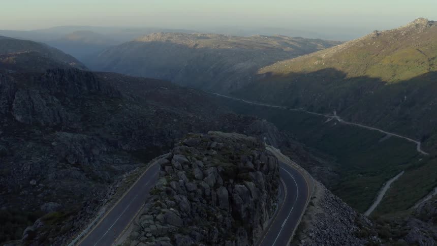 A curving freeway winds through the mountains with a distant mountain range glowing in the sunrise