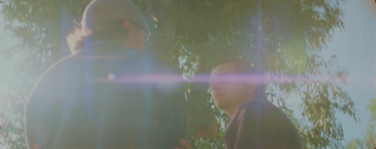 Two people have a conversation outdoors their figures partially obscured by a hazy dreamlike lens flare from the bright sunlight