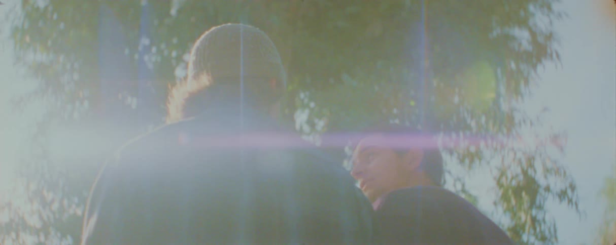 A young man has a conversation with another person outdoors as the scene is bathed in a hazy dream like sunlight