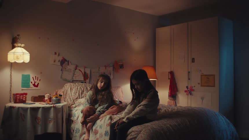 Two young girls sit sidebyside on the edge of a bed in a dimly lit cozy bedroom filled with childhood decorthey remain still and appear contemplative or somber while the soft glow of a bedside lamp illuminates the room