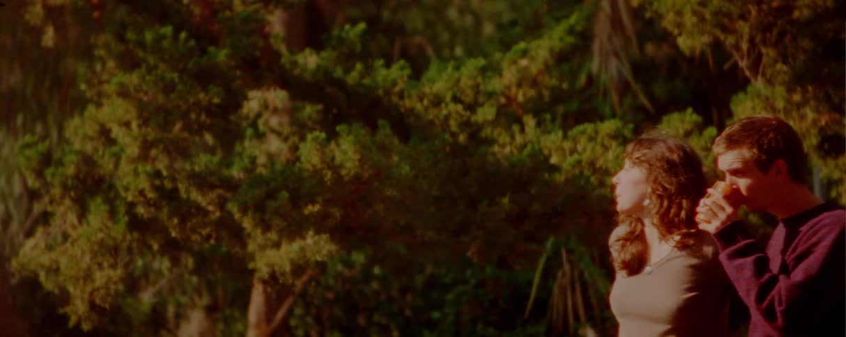 Teenager boy drinking a beverage from a disposable cup and walking together with a teenager girl next to another couple caressing each other at a park outdoors during sunset