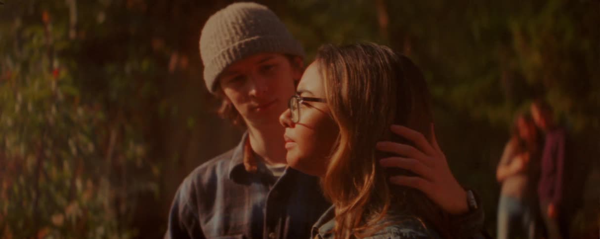 Younger man and woman are standing together with the man turned sidewayts looking at the girlhe strokes her hair and she takes a drink out of her cupthere is a couple in the background that are talking and touching each other as they stand together