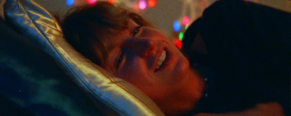 A young woman with red hair lies on a pillow and laughs joyfully during a conversation framed in a warm closeup with colorful outoffocus string lights decorating the dark background