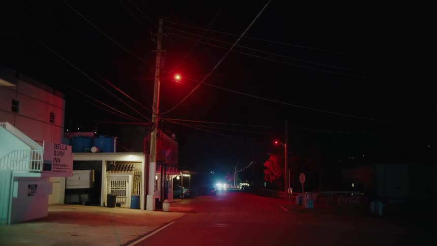 Street lights outside of buildings illuminate the street as automobile headlights glow in the distance