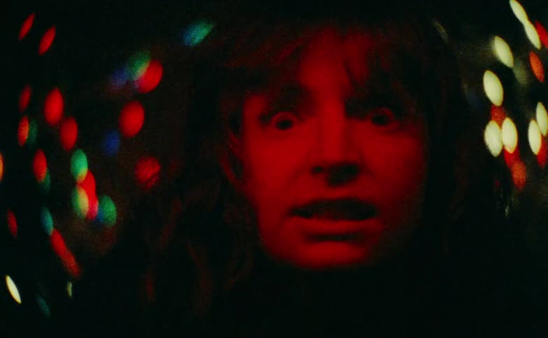 A woman with wide eyes and an open mouth looks directly into the camera with an expression of pure terror illuminated by a harsh red light against a dark background of blurry colorful bokeh lights