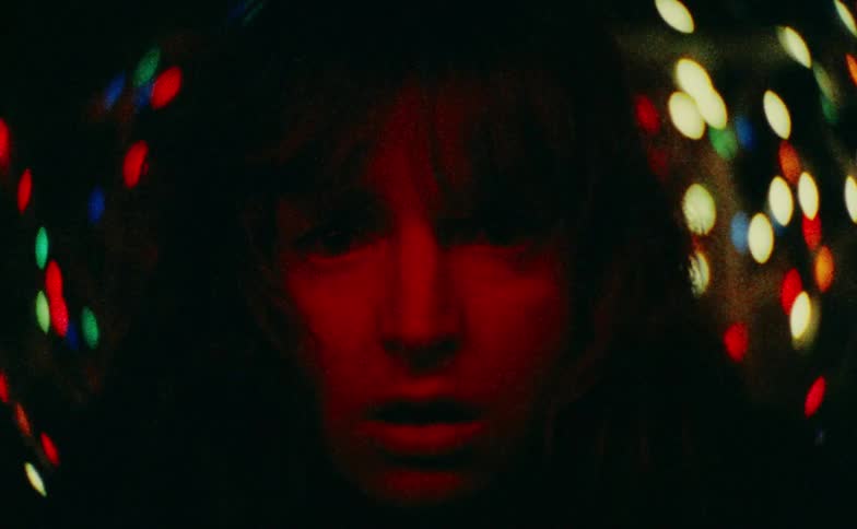 A woman with a shocked expression stares intensely forward as her face is bathed in a deep red lightin the final moments her mouth opens wide into a silent dramatic scream against a background of blurred colorful lights