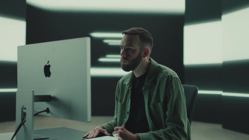 A bearded man sits at a desk in a modern studio pausing his writing to look away thoughtfully while positioned next to a large computer monitor and illuminated wall panels
