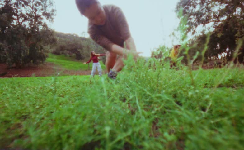 A woman in a brown shirt and tan shorts dances in a grassy field bending down doing expressive hand gestures while other people also dances in the distant background near trees and a sloping hillside