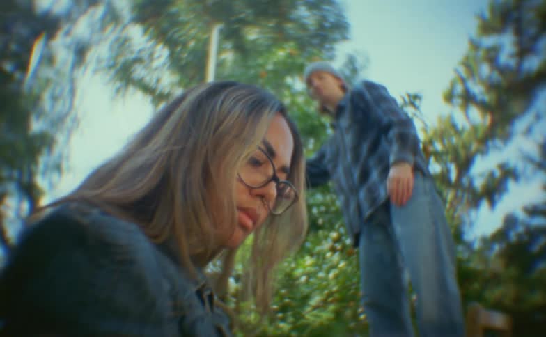 A woman with glasses looks downward with a somber expression in a lowangle closeup while a man in a flannel shirt and beanie approaches her from behind in a distorted outdoor setting