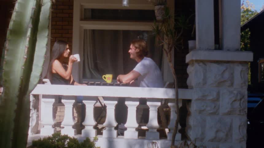 Two people seated on a porch talk while drinking coffee at a table