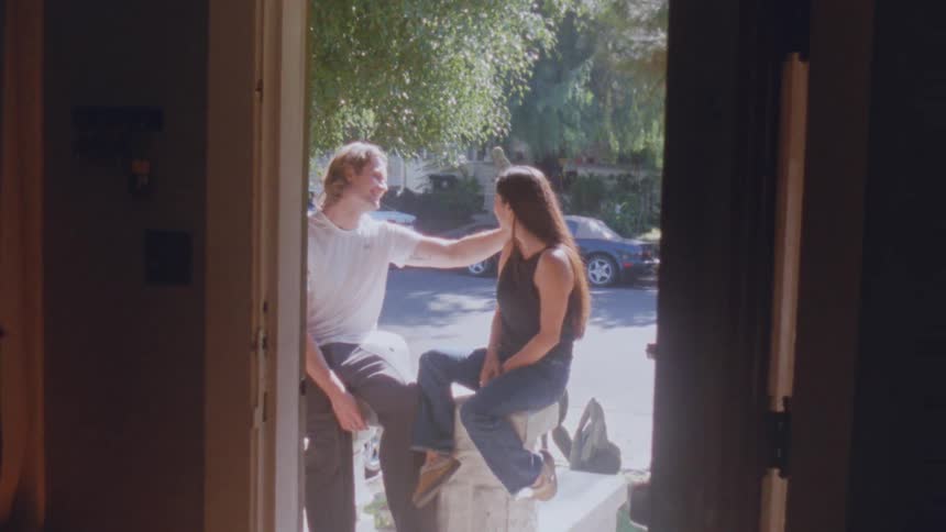 A young couple sits on a stool outdoors talking as the young man gently rubs the womans hair with his arm