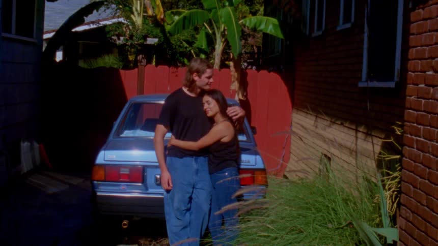 A man and woman stand in a sunny driveway by a blue car sharing an affectionate moment as he kisses her forehead and they embrace against a backdrop of a red fence and tropical foliage