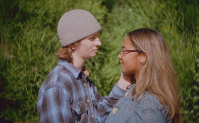 A young man in a flannel shirt and beanie gently cups a womans face as they gaze into each others eyes while standing outdoors in front of a tall green hedge during the day