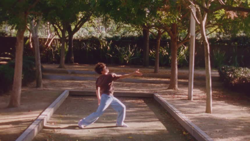 The boy in the brown shirt and jeans is dancing between the rows of trees and in front of the fence