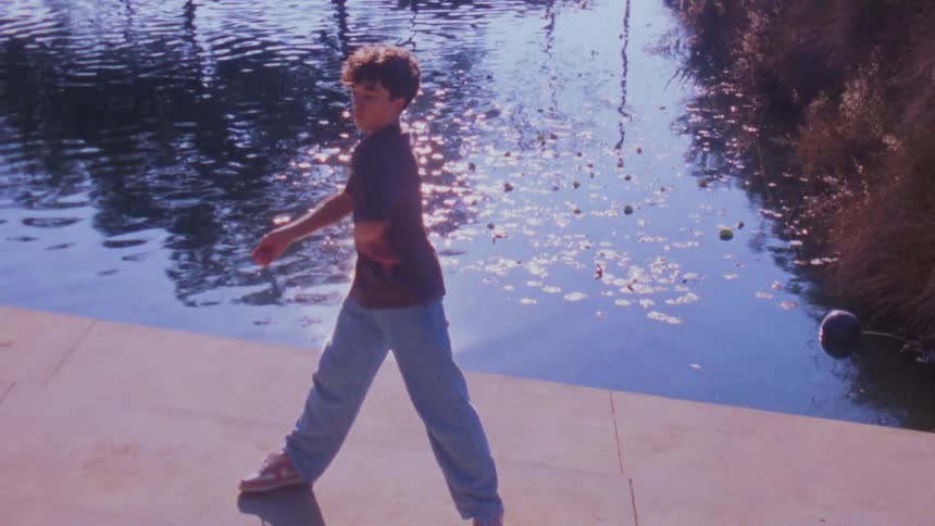 A teenage boy dances on a pavement by the riverbank showing off his moves as sunlight reflects across the surface of the water