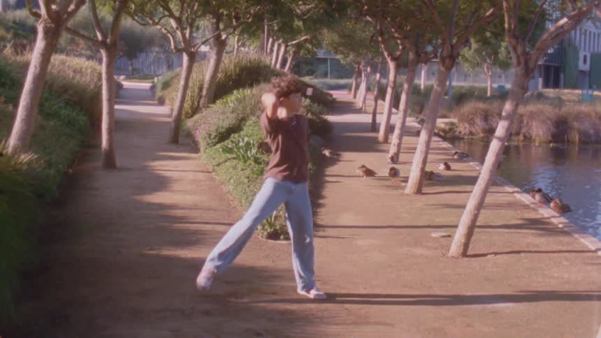 A young man in a tshirt and jeans performs an expressive rhythmic dance on a treelined dirt path next to a pond where several ducks are resting under the bright afternoon sun