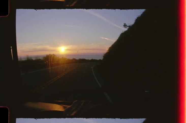 Windshield of a vehicle traveling on a highway surrounded by mountains and the sun on the horizon