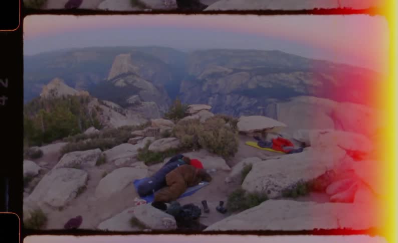 A group of people sleep in their sleeping bags atop a mountain at sunrise