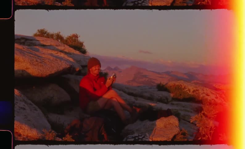 A person in a red jacket sits atop a rocky mountain peak during golden hour taking a photograph with a small camera before turning to smile