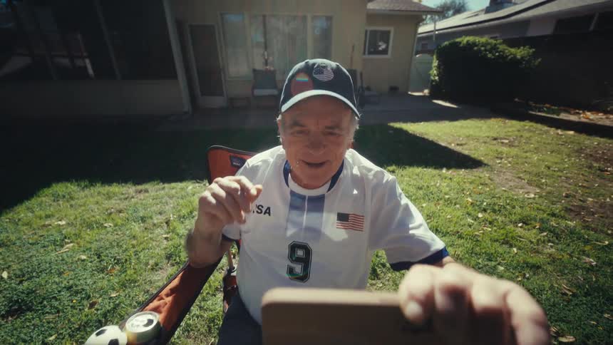 An elderly man excitedly watches sports on his smartphone in a backyard on sunny day