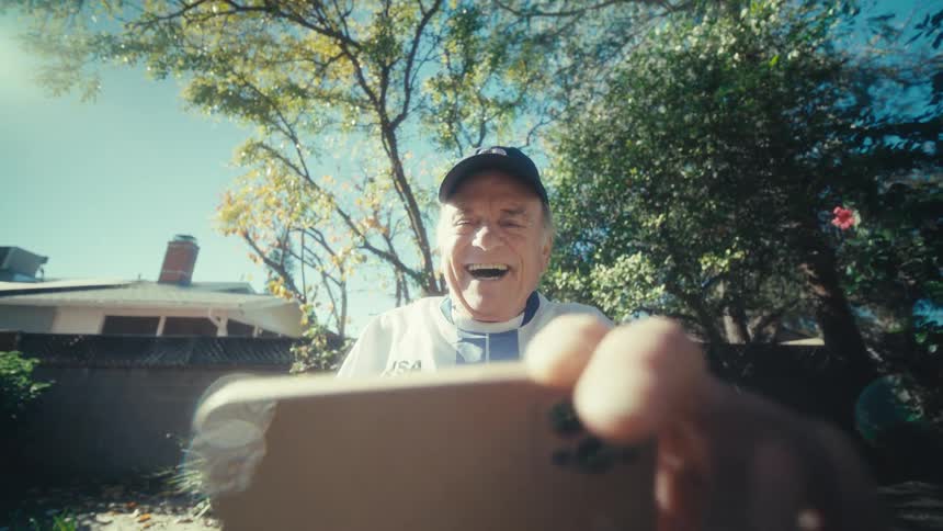 A joyful senior man in a usa jersey laughs heartily while holding up a smartphone in a sunny backyard surrounded by trees