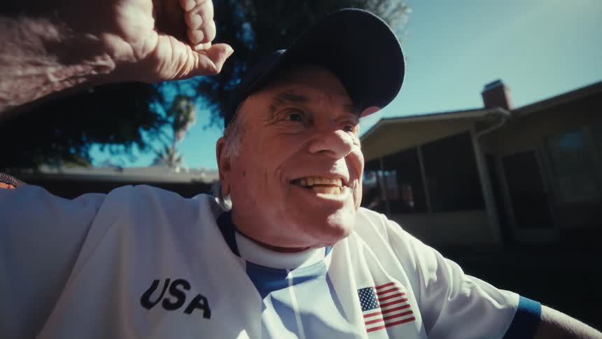 A senior man in a jersey excitedly watches sports on his smartphone in a sunny backyard