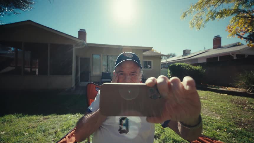 An elderly man cheers while watching a live sports match on his phone in a sunny backyard