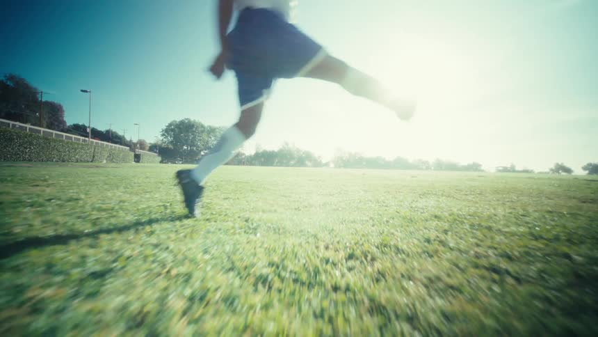 A man is runniing and kicks a soccer ball powerfully across dewbright green grass under a clear blue sky with sunlight flaring