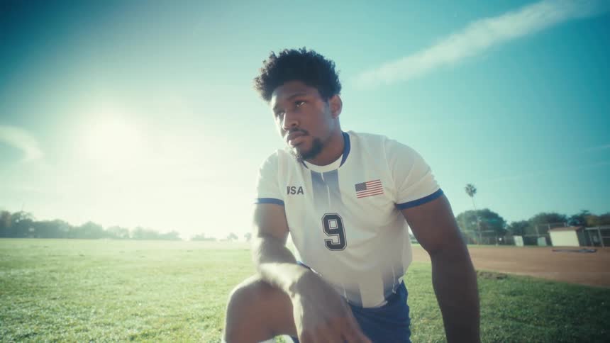 Soccer player kneeling to tie a cleat beside the ball on a grass field under a clear sky