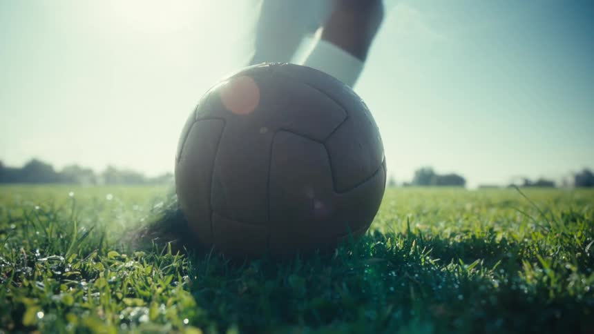 A soccer player kicks a ball on green grass under bright sunlight