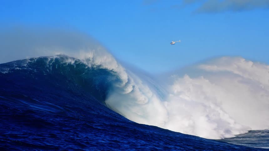 Helicopter hovers over the ocean as a large wave breaks under a bright blue sky