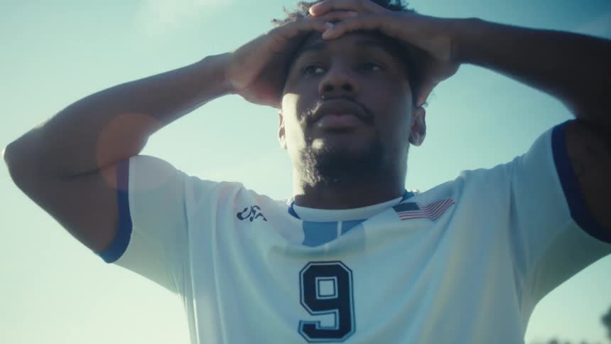 A man wearing a white usa soccer jersey with the number nine walks across a green grass field during a sunny day with bright lens flares