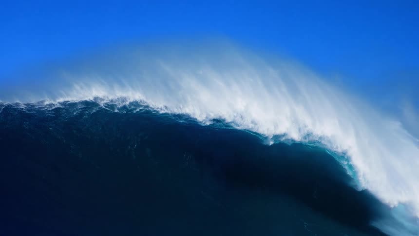 A massive blue ocean wave curls with white foam under a clear sky