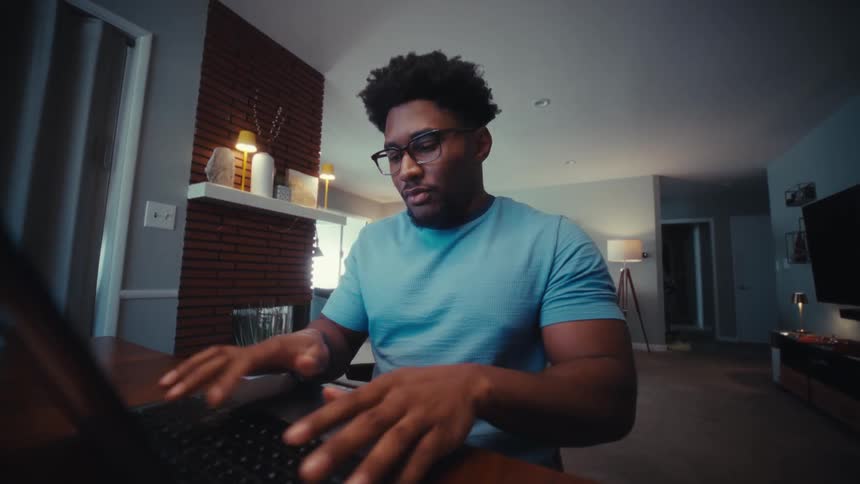 A man wearing a blue shirt and glasses sits at a wooden table in a living room and opens a laptop to start working during the evening