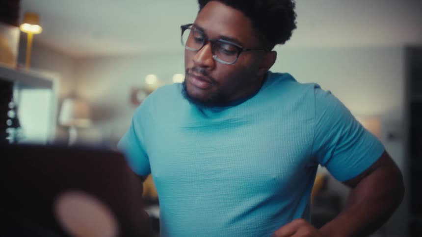A young man wearing glasses sits behind a desk in his living room at midday stretching his shoulders as he sets down a coffee mug and opens his computer lid