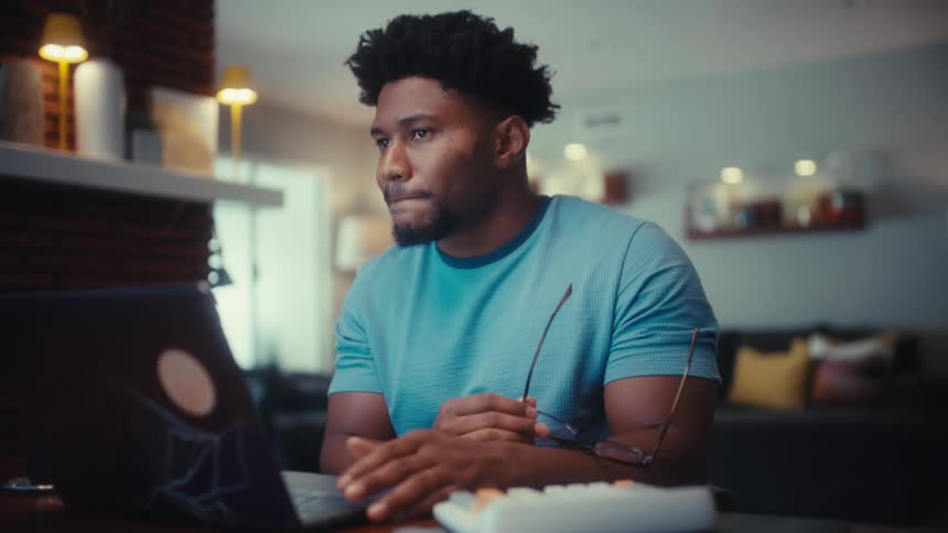 Thoughtful man sits in front of a laptop taking his glasses off and putting them back on