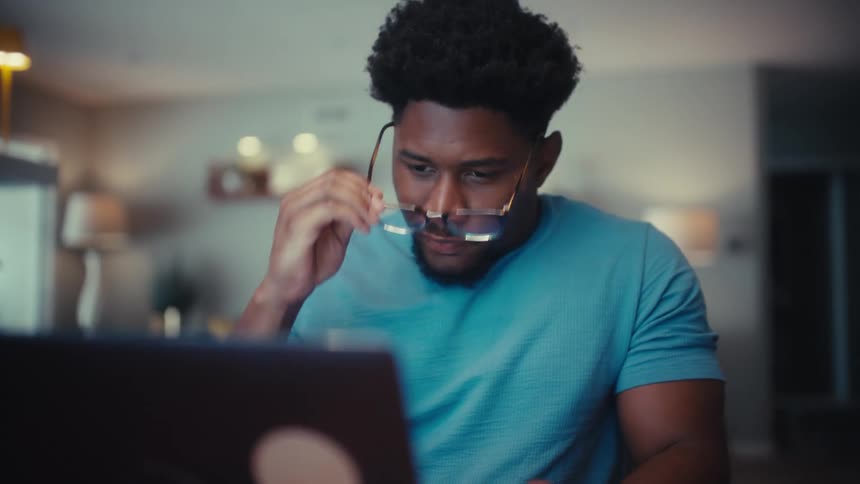 A man sits at a desk in a dimly lit room intensely focused on his laptop screen while typing midway through he removes his glasses to lean in for a closer look before putting them back on and continuing his work