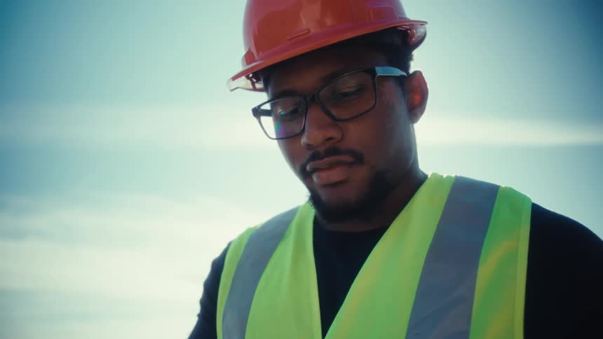 A worker wearing a helmet glasses and a green vest stands outdoors at midday reviewing information on his tablet under the bright sun