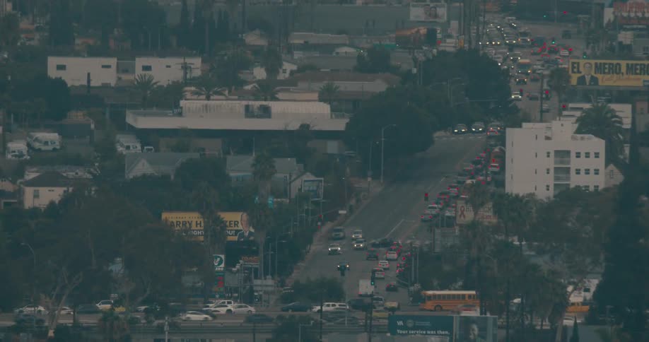 An aerial zoom moves from a wide shot of houses on a hazy hillside down toward a busy city street the lower shot reveals heavy traffic commercial buildings and billboards under a dim overcast sky