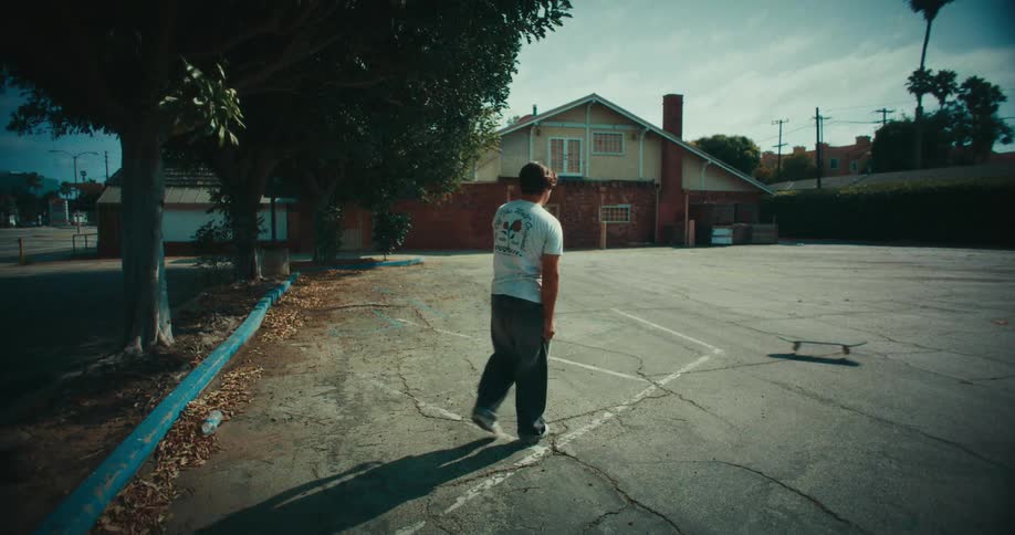 A man in a white tshirt and dark pants tosses a skateboard across an empty asphalt parking lot toward a brick building on a sunny day