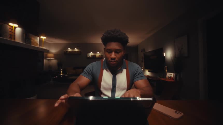A young black man using a tablet computer while sitting at a table indoors at night