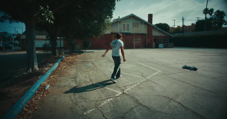 A person angrily throws a skateboard across an empty suburban parking lot