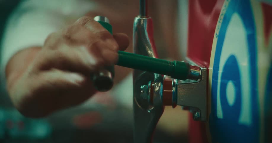 A persons hand uses a green ttool to adjust the kingpin nut on a silver skateboard truck the tool is rotated clockwise to tighten the truck against the colorful red and blue deck