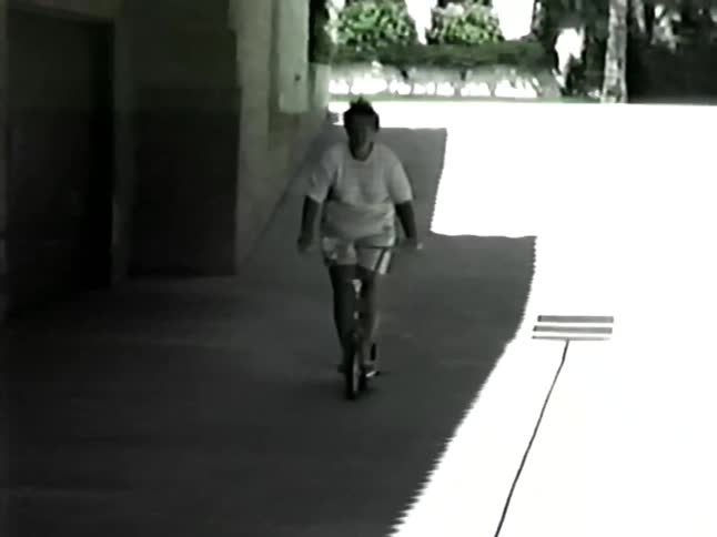 A woman in a white tshirt and glasses rides a bicycle with pink streamers down a sunny driveway moving from a bright outdoor area into the shade of a building toward the camera