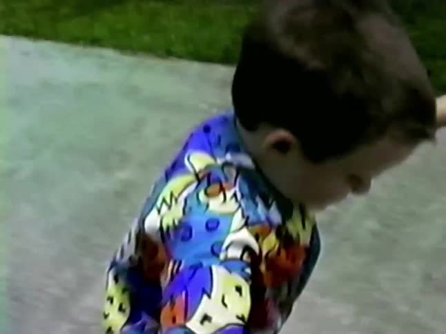 A young boy in a colorful patterned shirt smiles and laughs while riding a white scooter along a residential sidewalk during the day passing by a lawn and a parked car while other children walk nearby in the background
