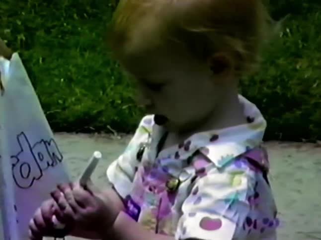 A woman assists a boy with a sign on his scooter while a toddler girl in pink overalls stands nearby on a sidewalk looking at a small toy in her hands during a sunny day outdoors