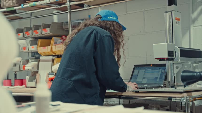 Person wearing a blue cap and dark jacket works on a laptop at a workshop bench with equipment and organized boxes in the background