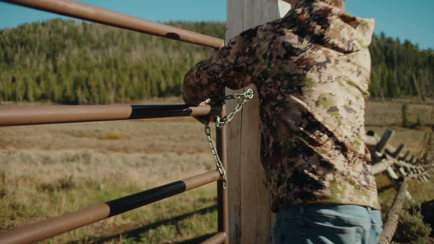 A man wearing a camouflage jacket and a baseball cap walks toward a metal ranch gate he reaches out to unhook a chain securing the gate to a thick wooden post in a rural mountainous setting